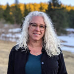 A woman with long white hair standing in front of trees.